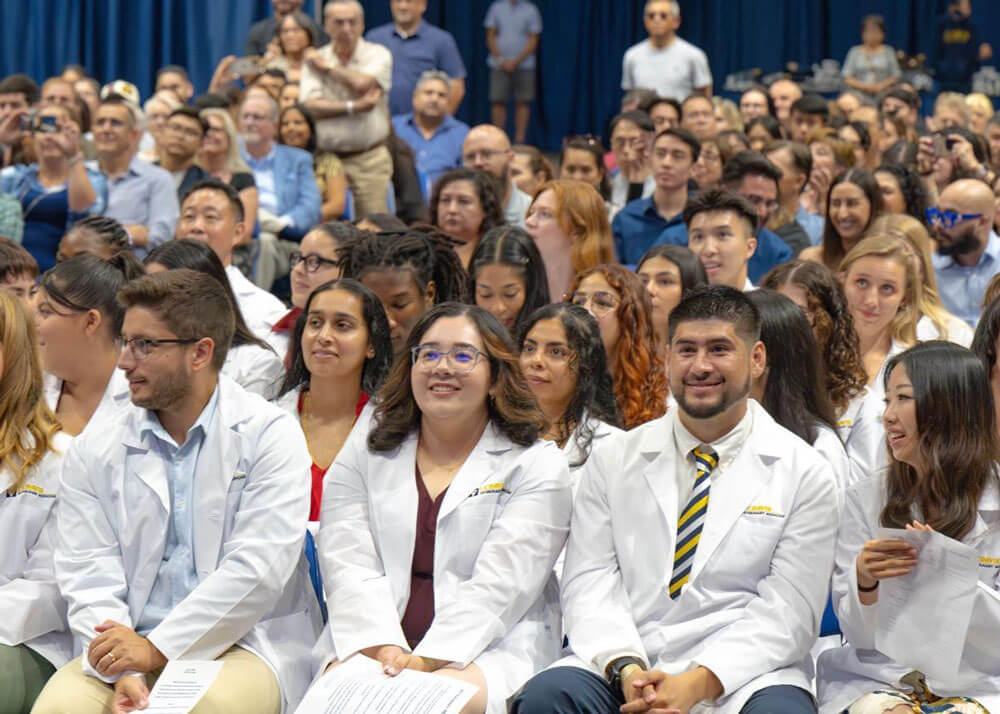 Imagining the future Veterinary Education Pavilion. Veterinary students wearing white lab coats while smiling and  seated in an auditorium. 