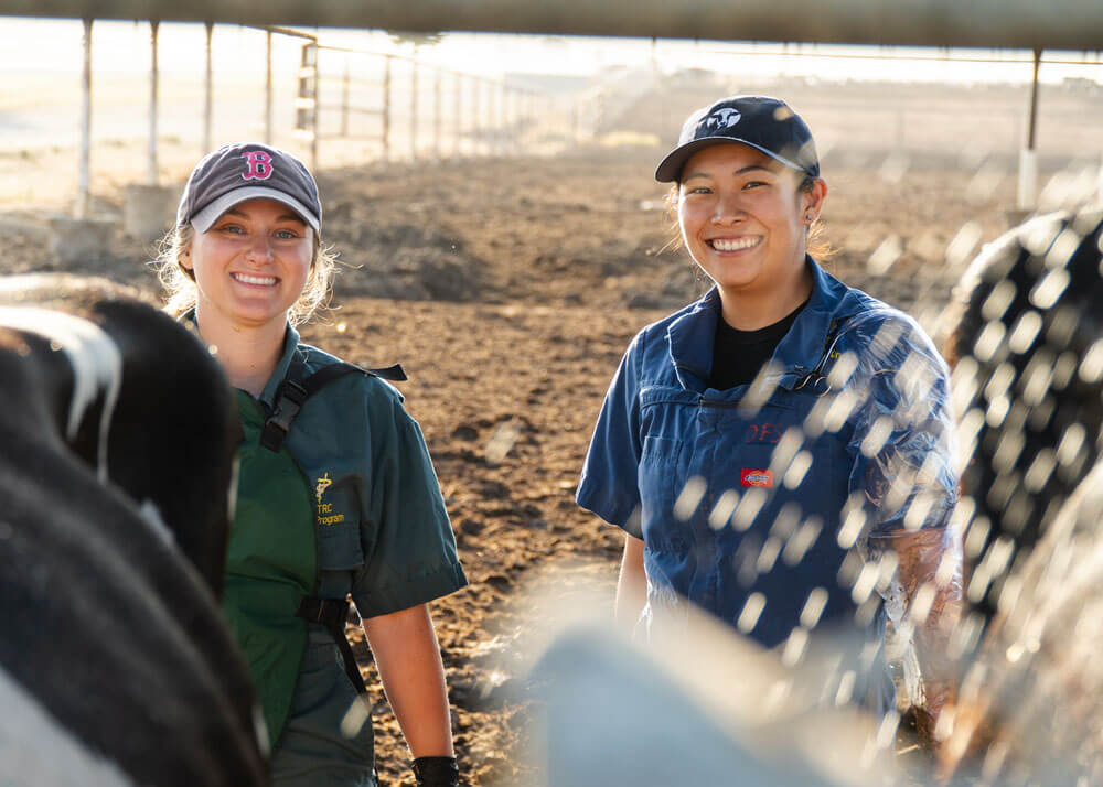 Two female veterinary students wearing baseball caps standing together in a large animal enclosure while smiling at the camera.