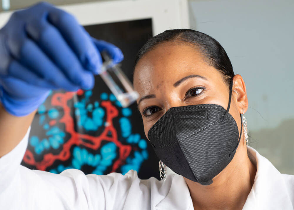 Close-up of a woman wearing a white lab coat, purple gloves and a face mask looking at some clear liquid in a vial. She is involved in discovery and innovation.