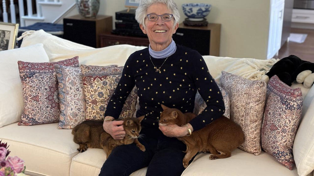 An older woman sits on a couch smiling at the camera as she cuddles two cats on her lap.