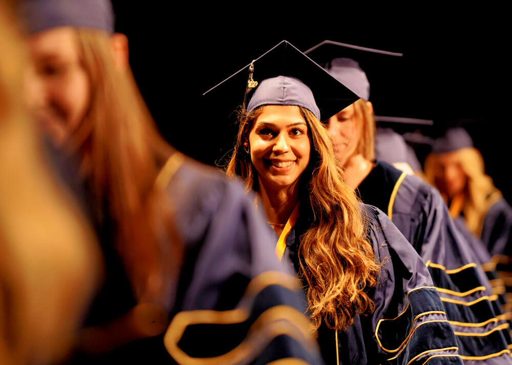 Graduating veterinary students wearing their caps and gowns, with a focus on one female smiling and walking in line with the others.