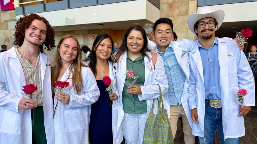 Six veterinary students wearing white lab coats and holding individual roses stand together smiling.