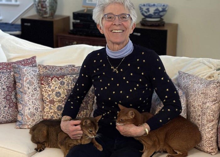 Lin Zucconi sits on a couch smiling and cuddling her two Abyssinian cats.