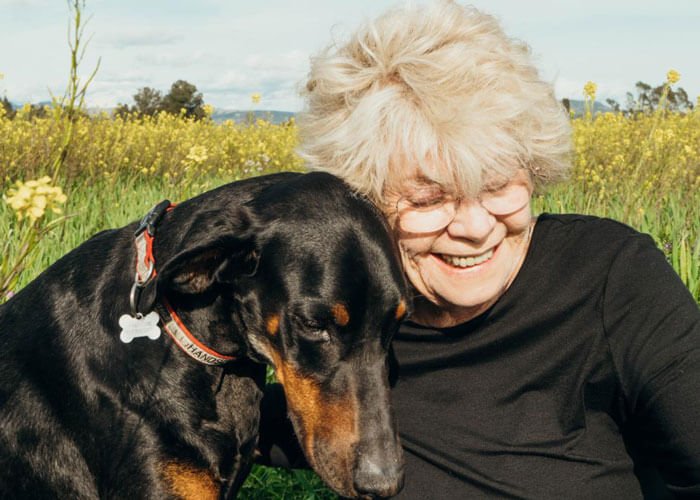 Judith Friedman smiles cuddles with her dog, a Doberman pinscher, which sits next to her.